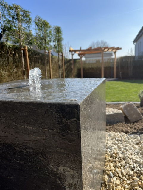 Water feature as a natural stone cube with gravel bed, pergola and lawn behind